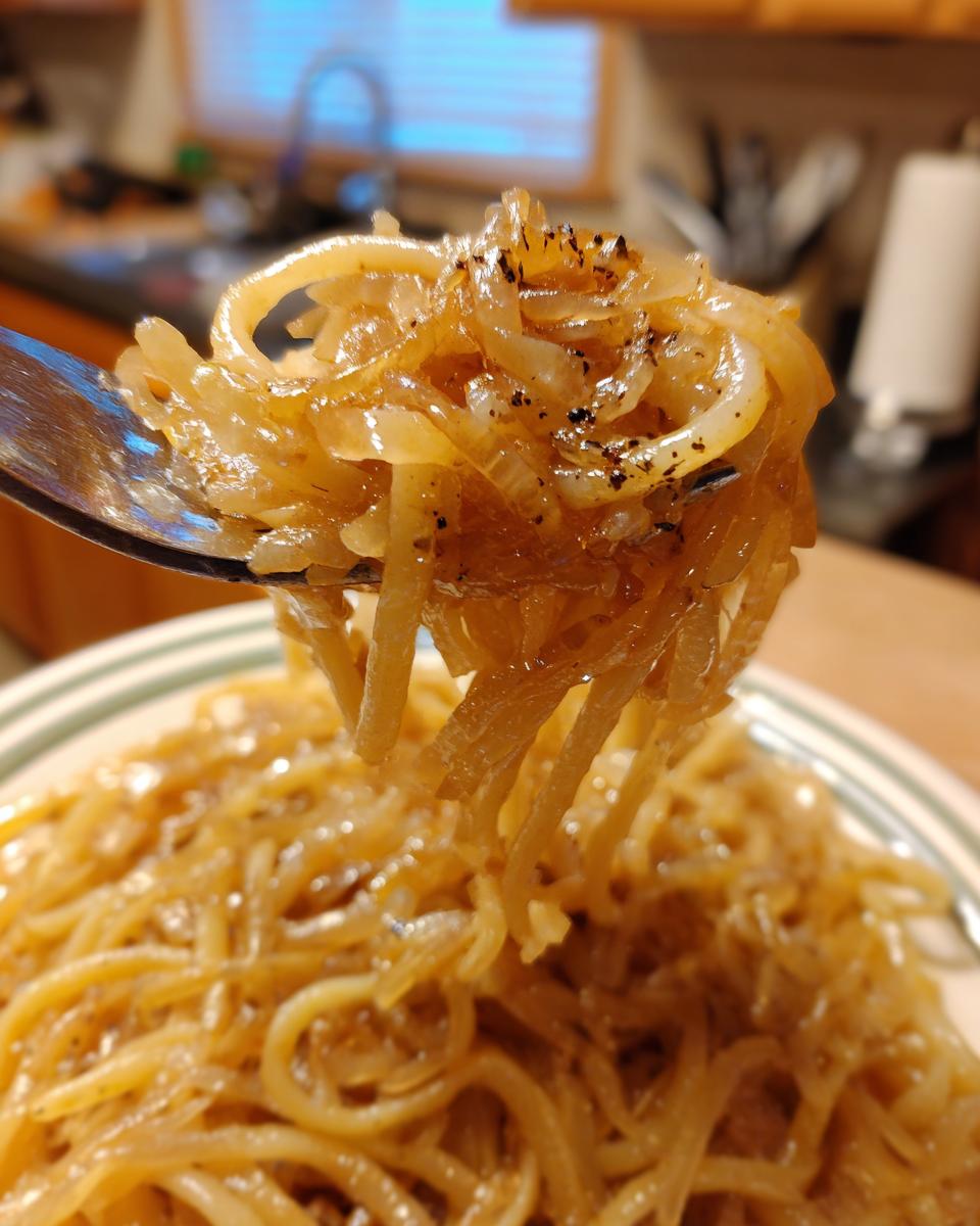 Close-up of a fork lifting pasta coated in rich caramelized onion pasta, seasoned with black pepper.