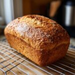 A golden brown, freshly baked loaf of whole wheat bread resting on a wire cooling rack.