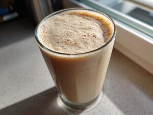 Close-up of a tall glass filled with a thick, frothy peanut butter banana smoothie, sitting on a light countertop near a window.