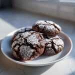 A close-up of four fudgy chocolate crinkle cookies dusted heavily with powdered sugar on a white plate.