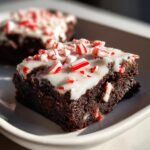 A close-up of a fudgy peppermint brownie square topped with white glaze and crushed candy canes.