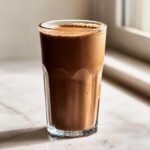 A tall glass filled with a thick, brown Coffee Smoothie sitting on a marble counter near a window.