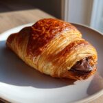 A freshly baked, flaky chocolate croissant with melted chocolate oozing out, sitting on a light plate.