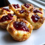 Close-up of several golden, flaky puff pastry brie bites topped with melted brie and a dollop of red jam.