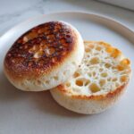Close-up of a homemade english muffin split in half, showing the signature open, craggy interior and toasted, bubbly exterior.