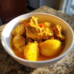 A close-up of vibrant yellow Jamaican curry chicken and potatoes served in a white bowl.
