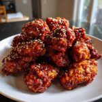 A pile of glossy, dark red korean fried chicken pieces topped with white sesame seeds on a white plate.