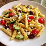 A close-up view of a serving of mediterranean pasta salad featuring penne, cucumber slices, cherry tomatoes, olives, and feta cheese.