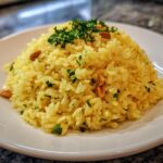 A mound of vibrant yellow mediterranean rice garnished with fresh parsley and toasted pine nuts on a white plate.