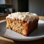 A close-up of a moist slice of applesauce cake topped with thick cream cheese frosting, sitting on a gray plate.