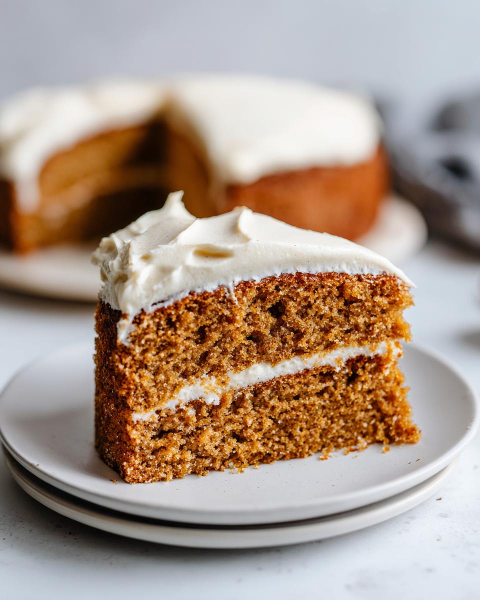 Close-up of a thick slice of moist applesauce cake layered with white frosting, with the rest of the cake blurred in the background.