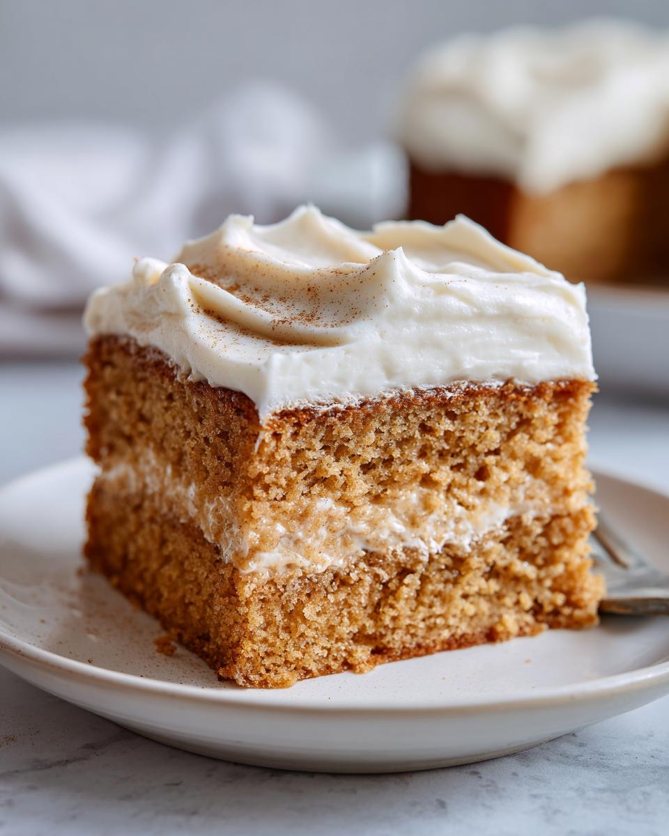 Close-up of a square slice of moist applesauce cake with a layer of filling and thick cream cheese frosting dusted with cinnamon.