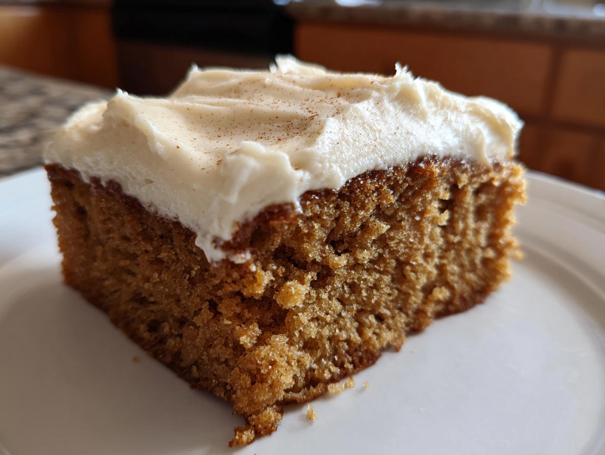 A close-up of a moist slice of applesauce cake topped with thick, white cream cheese frosting dusted with cinnamon.