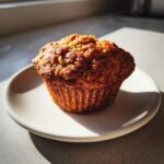 A single, golden-brown, moist bran muffin sitting on a small white plate, highlighted by bright sunlight.