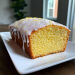 A close-up of a freshly baked lemon loaf cake, topped with thick white glaze and lemon zest.
