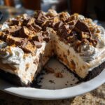 A rich, no-bake butterfinger pie with a slice removed, showing the creamy filling and chocolate crust, topped with whipped cream and candy pieces.