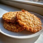 Three golden brown, crispy oatmeal lace cookies stacked on a white plate near a sunlit window.