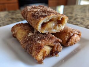 Three golden-brown peach cobbler egg rolls dusted heavily with cinnamon sugar, one cut open to show the filling.