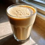 A tall glass filled with a thick, frothy peanut butter banana smoothie, catching sunlight near a window.