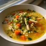 Close-up of a white bowl filled with golden penicillin soup containing shredded chicken, carrots, celery, and Israeli couscous.