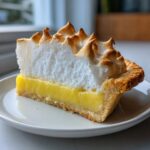 A perfect slice of lemon meringue pie showing thick, torched meringue over bright yellow curd in a flaky crust.