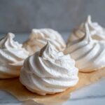 Close-up of several perfectly piped, white meringue cookies resting on brown parchment paper.