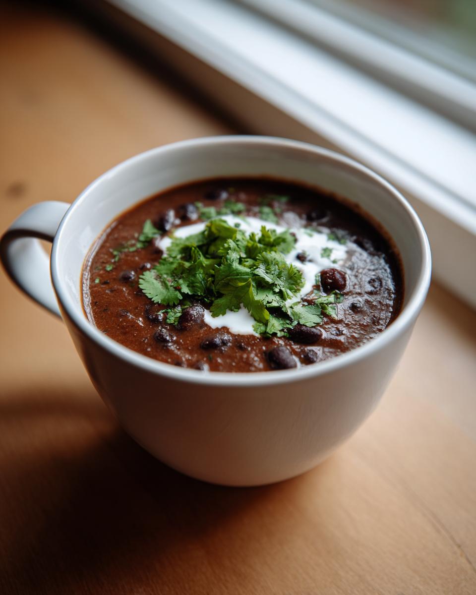 A close-up of a mug filled with rich, dark black bean soup, topped with sour cream and fresh cilantro.