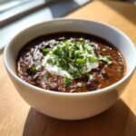 A close-up of a bowl of thick, dark black bean soup topped with a dollop of sour cream and fresh cilantro.