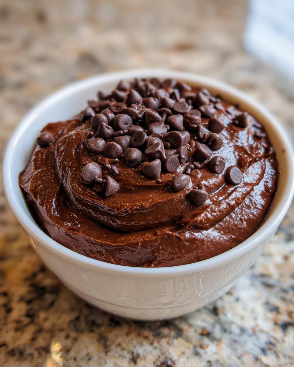 A close-up of rich, thick brownie batter dip topped generously with chocolate chips in a white bowl.