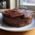 A round cake generously covered in thick, dark chocolate frosting, sitting on a white plate.