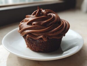 A perfectly piped swirl of rich chocolate frosting topping a dark chocolate cupcake on a white saucer.