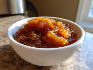 Close-up of rich, dark brown spiced applesauce served in a small white bowl, glistening in the sunlight.