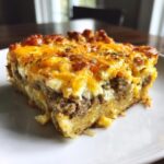 A thick slice of savory egg bake featuring layers of hash browns, sausage, and melted cheddar cheese.