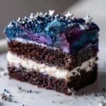 Close-up of a slice of dark chocolate capricorn cake layered with white cream and topped with galaxy-themed blue and purple frosting and silver sprinkles.