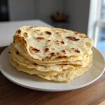 A tall stack of freshly cooked, slightly charred corn tortillas resting on a white plate.