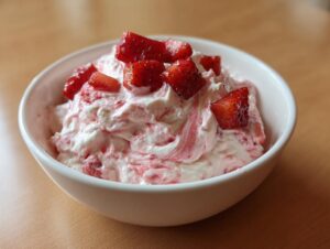 A close-up of creamy, swirled strawberry cheesecake dip topped with fresh strawberry chunks in a white bowl.