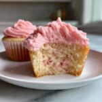 Close-up of a strawberry cupcakes cut in half showing the moist crumb with strawberry pieces and pink frosting.