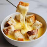 A piece of crusty bread is lifted from a white bowl of melted cheese fondue, showing a long, stretchy cheese pull.