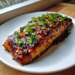 A perfectly cooked fillet of teriyaki salmon, glistening with dark glaze, topped with sesame seeds and chopped green onions.