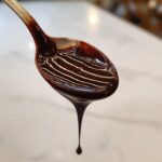 Close-up of thick, glossy chocolate sauce recipe dripping from a silver spoon.