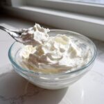 A spoonful of thick, bright white royal icing recipe mixture being lifted from a clear glass bowl.