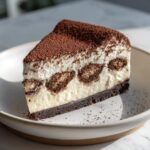 A perfect slice of tiramisu cheesecake featuring layers of cream, soaked ladyfingers, and a dark crust, heavily dusted with cocoa powder.