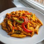 A close-up of creamy, orange-hued rasta pasta mixed with chunks of chicken and colorful bell pepper strips.