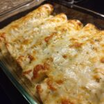 Close-up of baked white chicken enchiladas covered in bubbling, melted white cheese and herbs in a glass dish.