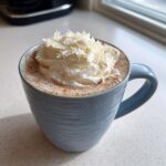 A mug of rich white hot chocolate topped with whipped cream and white chocolate shavings.