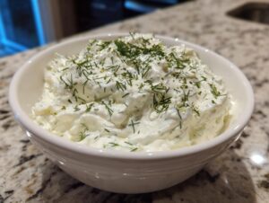 A close-up of creamy, white dill dip topped generously with fresh dill, served in a small white bowl.
