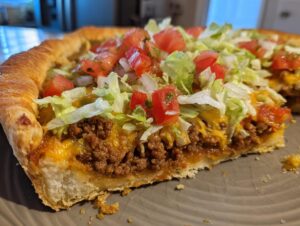 A thick slice of amazing taco pizza showing seasoned ground beef, melted cheese, lettuce, and diced tomatoes.