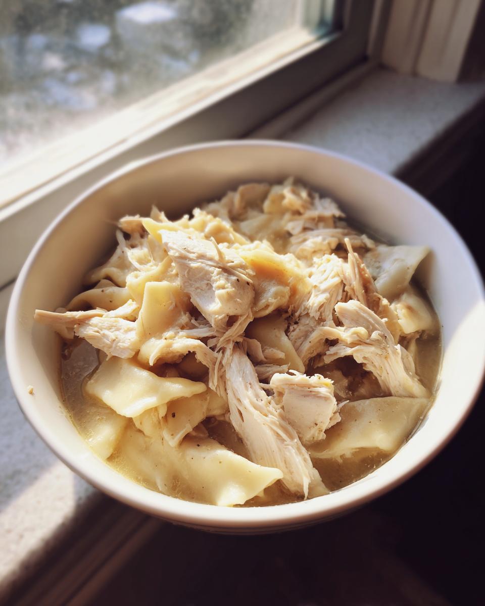 A close-up of a bowl filled with rich chicken and dumpling soup, featuring shredded chicken and flat noodles.