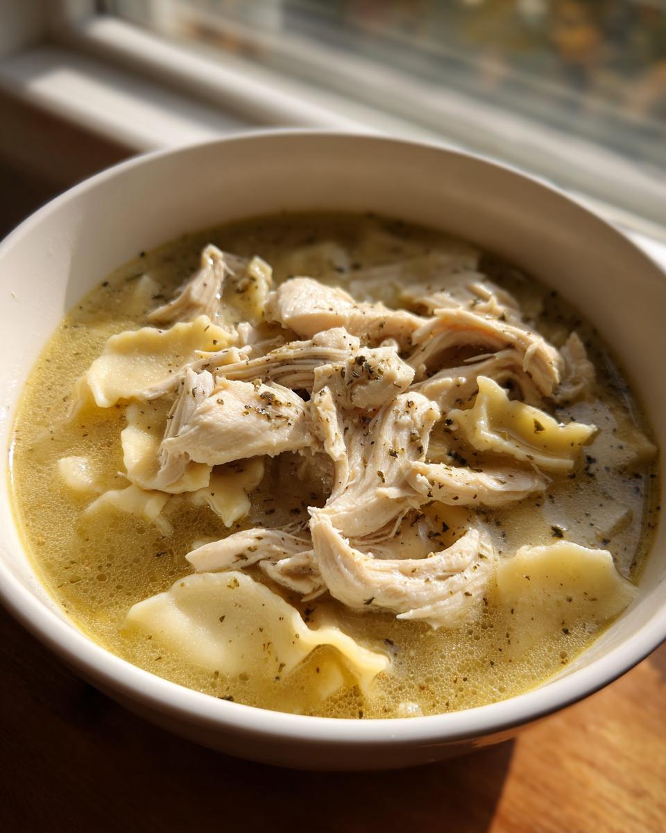 Close-up of a bowl of rich chicken and dumpling soup topped with shredded chicken and herbs.