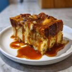 A decadent square slice of bread pudding with bourbon sauce generously drizzled over the top and pooling on the white plate.